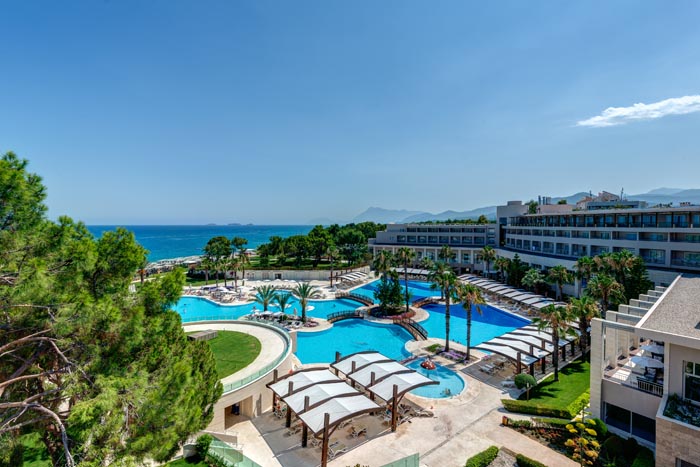Rixos Premium Tekirova panorāmas skats uz kūrorta teritoriju, baseiniem un Vidusjūru — apkārtnē zaļojošas priedes un kalnu silueti