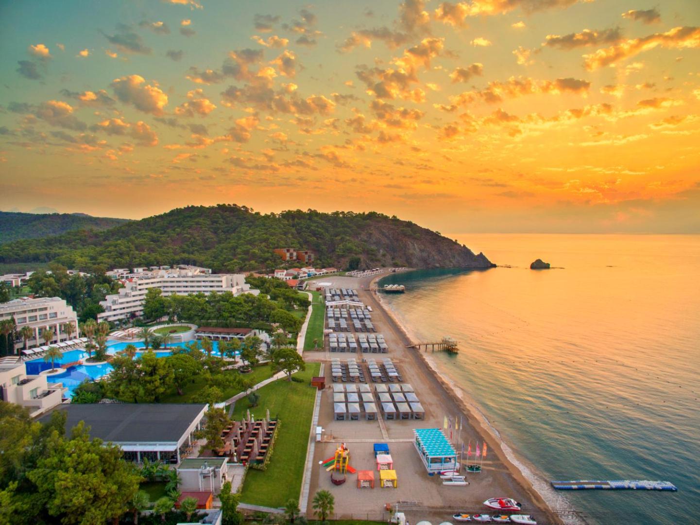Rixos Premium Tekirova panorāmas skats saulrietā — viesnīcas komplekss, baseini un plaša pludmale Kemeras piekrastē Turcijā