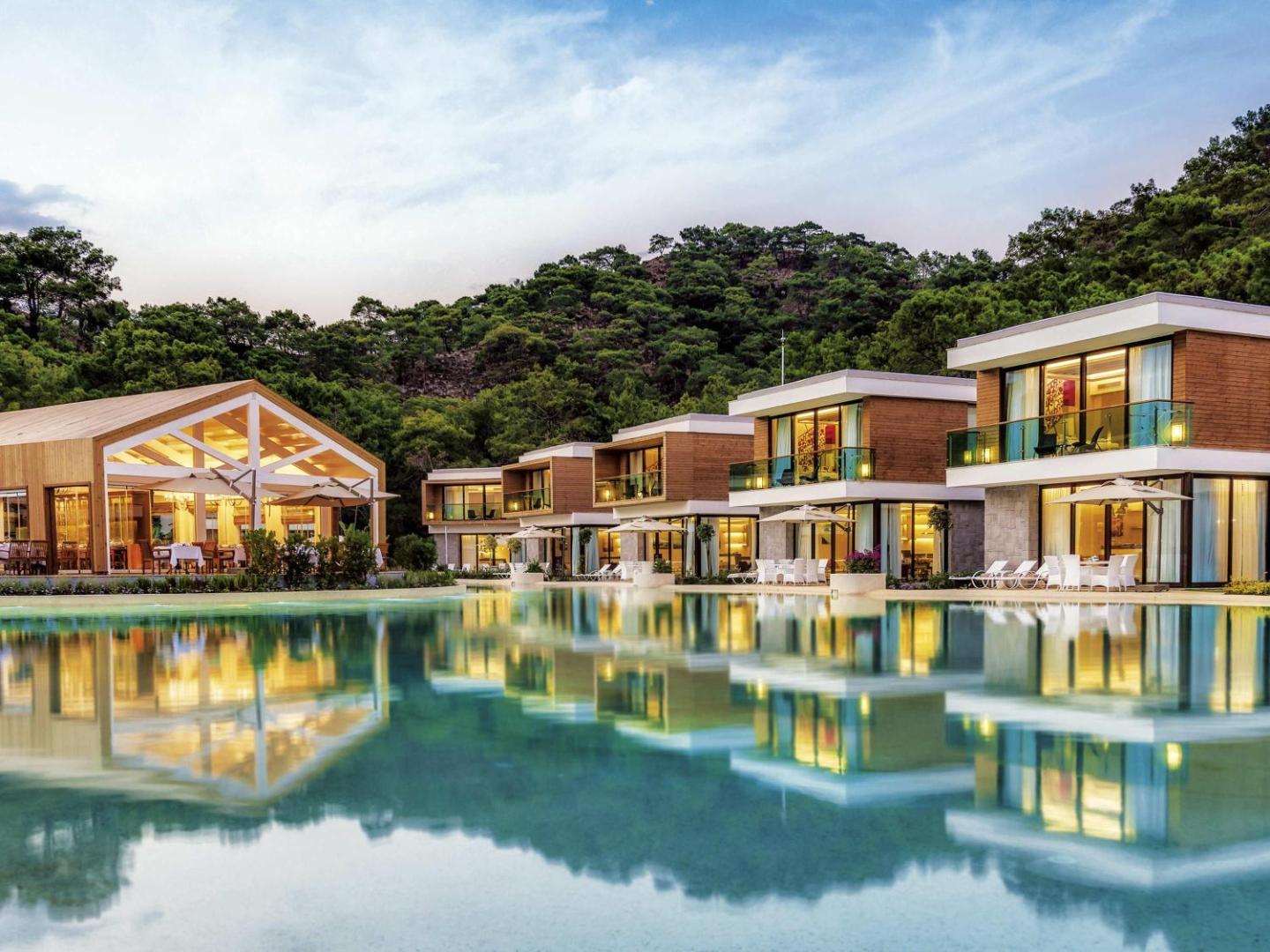 Rixos Premium Tekirova modernie divstāvu bungalo pie baseina vakara apgaismojumā ar priežu mežu fonā