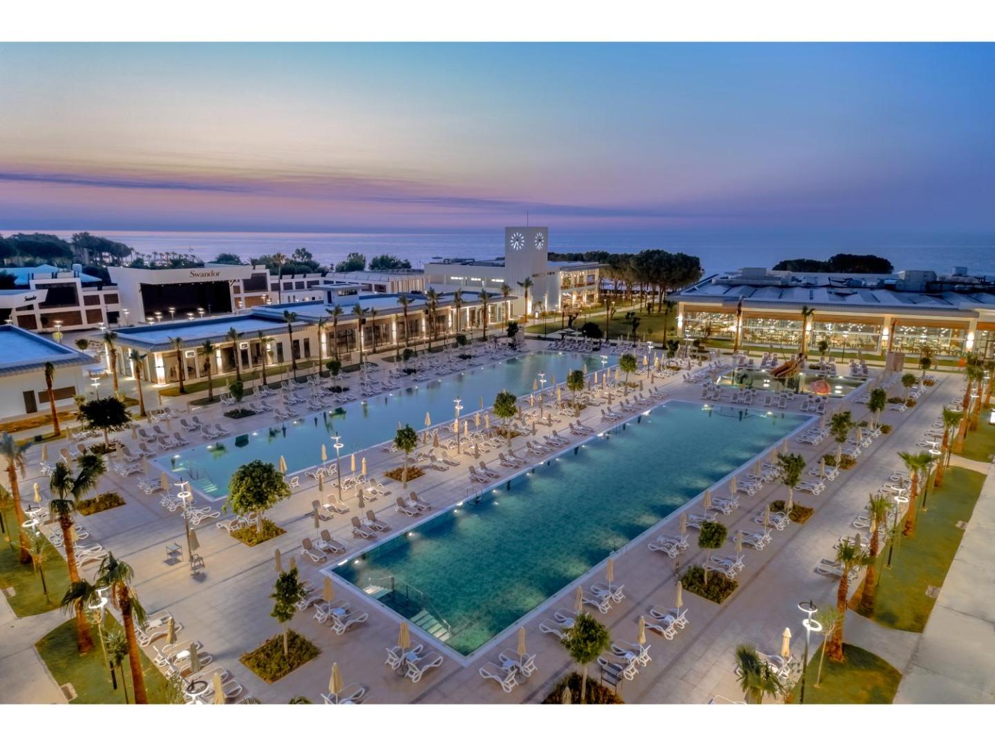 Swandor Hotels & Resorts Kemer panorāmas skats krēslas stundā — lieli āra baseini, izgaismota teritorija un Vidusjūra fonā Turcijā