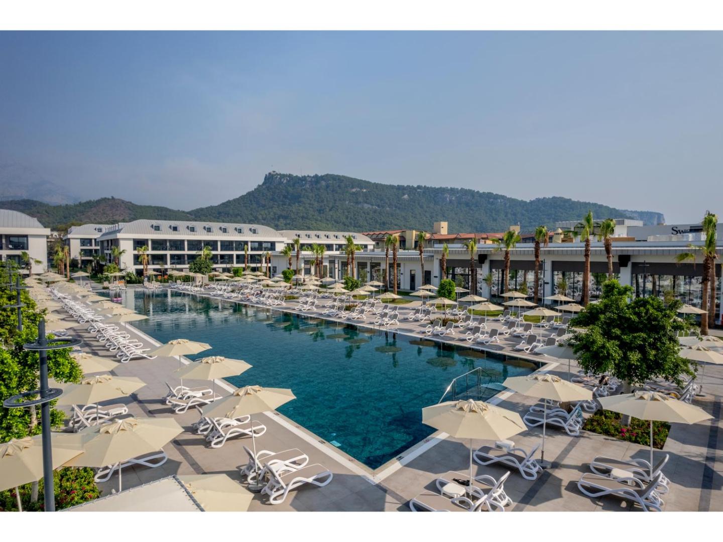 Swandor Hotels & Resorts galvenais baseins dienas laikā — balti saulessargi, sauļošanās krēsli un iespaidīgs skats uz kalniem