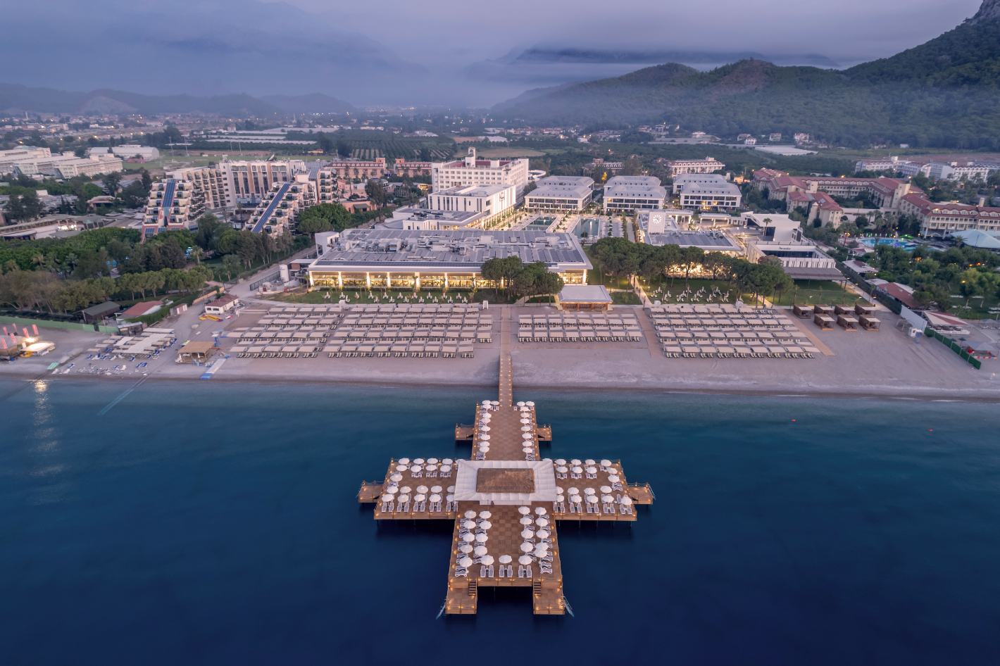 Swandor Hotels & Resorts Kemer panorāmas skats no putna lidojuma uz viesnīcas kompleksu, privāto pludmali un koka piestātni Vidusjūras krastā
