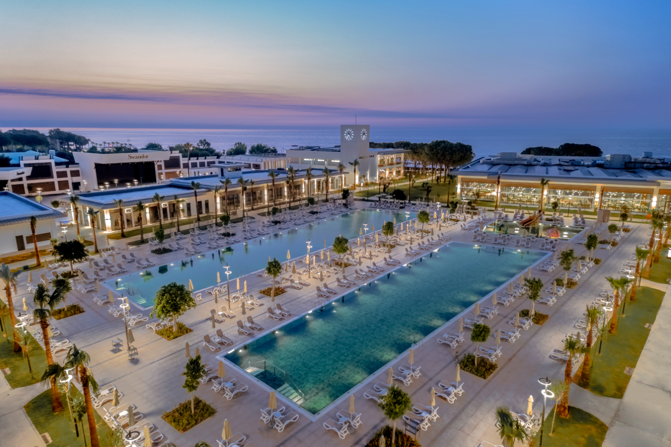 Swandor Hotels & Resorts plašā baseinu zona saulrietā — skats no augšas uz sauļošanās krēsliem, palmām un viesnīcas korpusiem