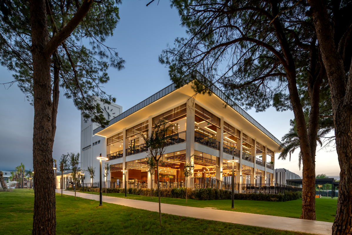 Swandor Hotels & Resorts Kemer modernā restorāna ēka vakarā ar izgaismotām terasēm, priežu ieskautu zālāju un pastaigu taku