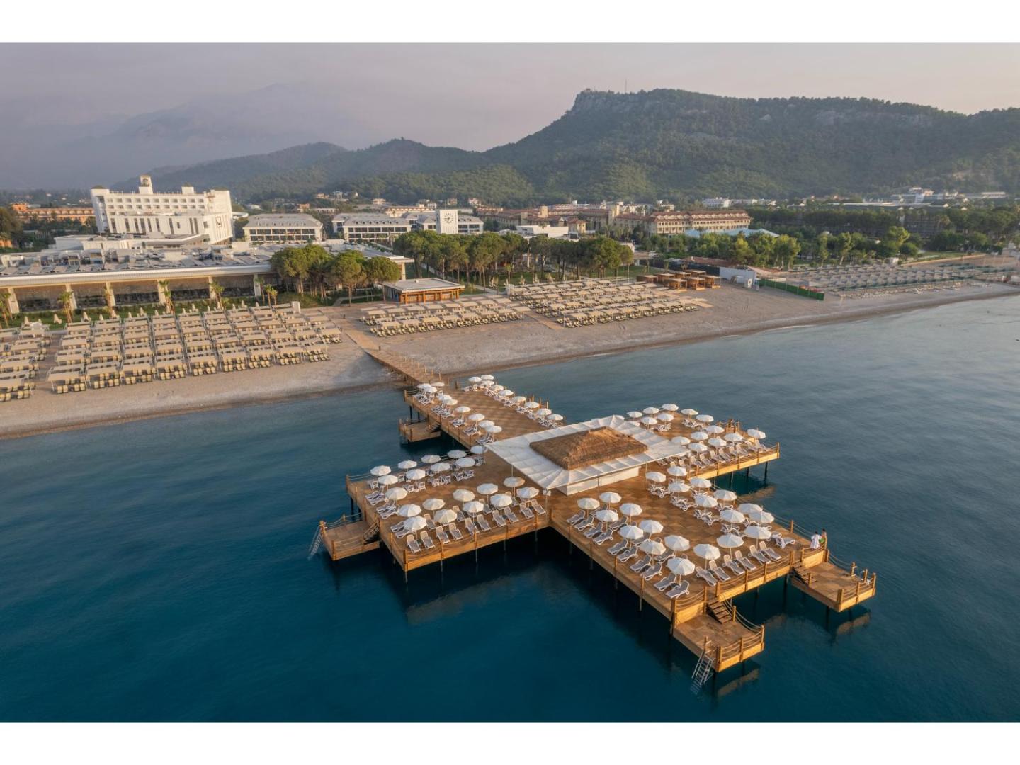 Swandor Hotels & Resorts Kemer koka piestātnes tuvplāns ar baltiem saulessargiem un skatu uz piekrasti un kalniem saullēktā