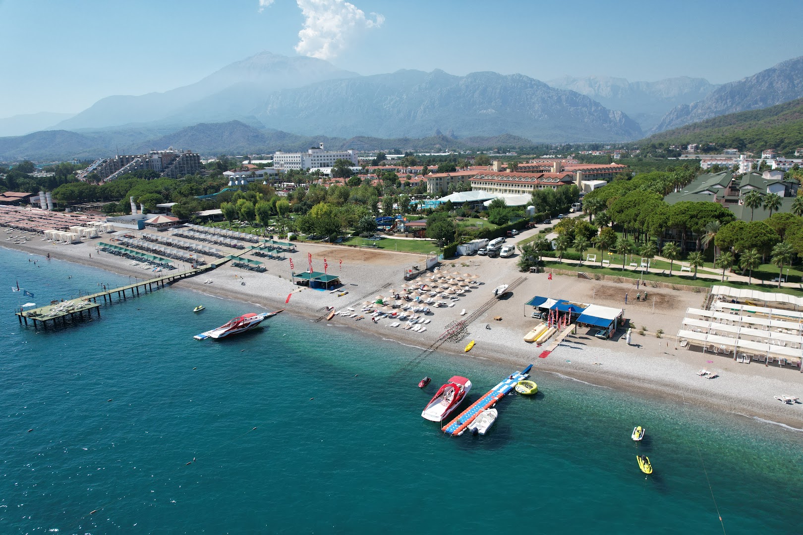 Swandor Hotels & Resorts Kemer pludmales zona ar saulessargiem, ūdens sporta centru un kalnu ainavu fonā saulainā dienā