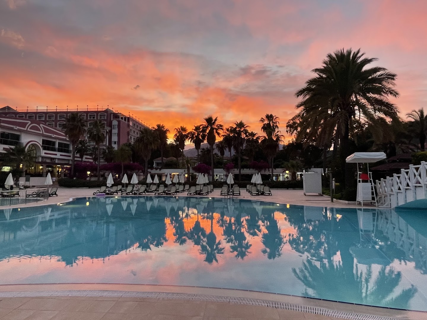 Swandor Hotels & Resorts baseins saulrieta laikā ar palmu atspulgu ūdenī — rozā debesis un kalnu siluets fonā