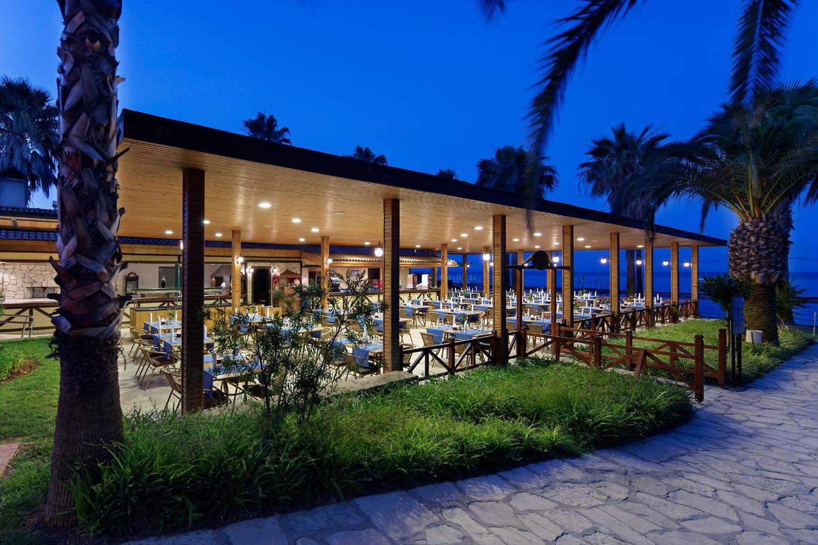 Meryan Hotel āra restorāna terase vakarā ar galdiņiem zem koka nojumes un palmām dārzā pie jūras