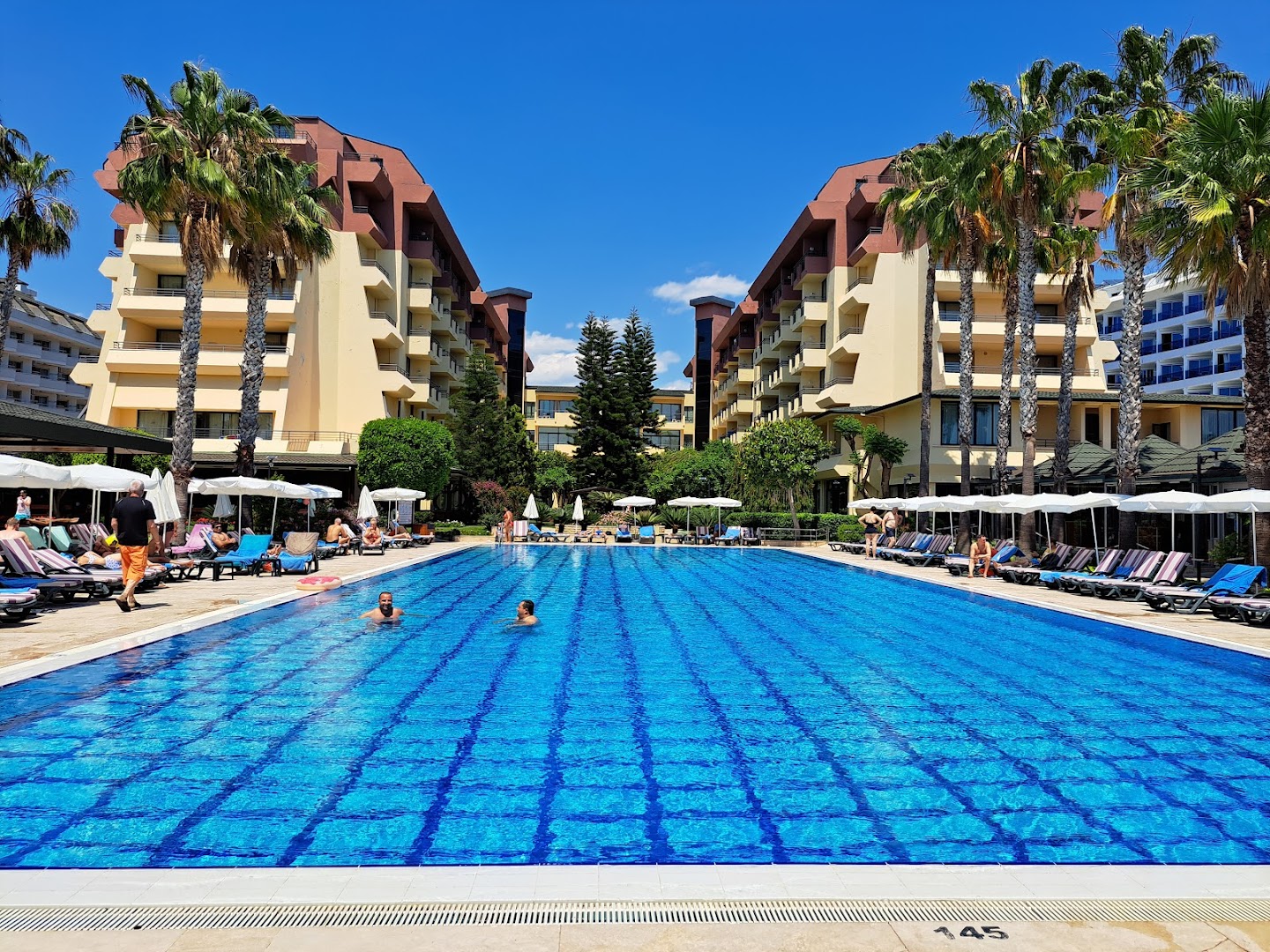 Meryan Hotel centrālais peldbaseins starp viesnīcas korpusiem ar sauļošanās krēsliem, saulessargiem un palmām zem skaidrām debesīm.