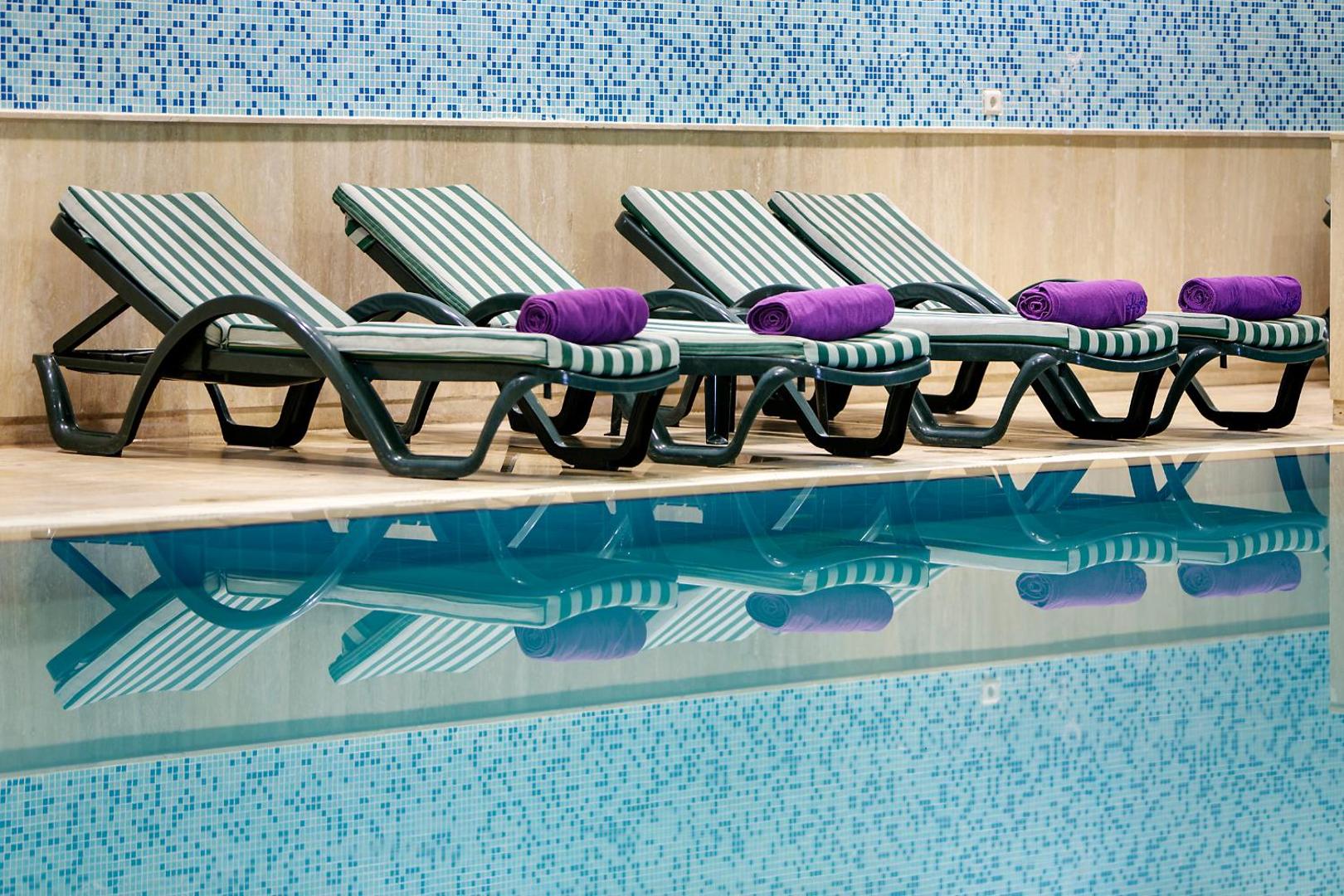 Meryan Hotel iekštelpu baseins ar svītrainiem zviļņiem un violetiem dvieļiem — atpūtas zona pie ūdens ar zilu mozaīkas sienu