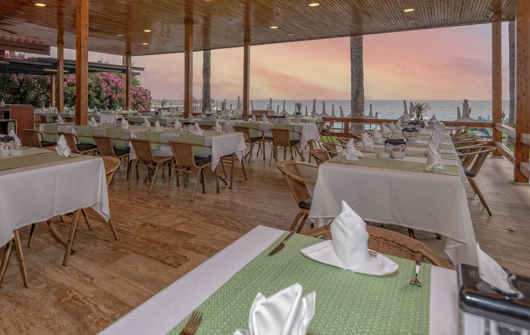 Meryan Hotel restorāns ar skatu uz jūru, klātiem galdiem un koka griestiem saulrieta debesu fonā