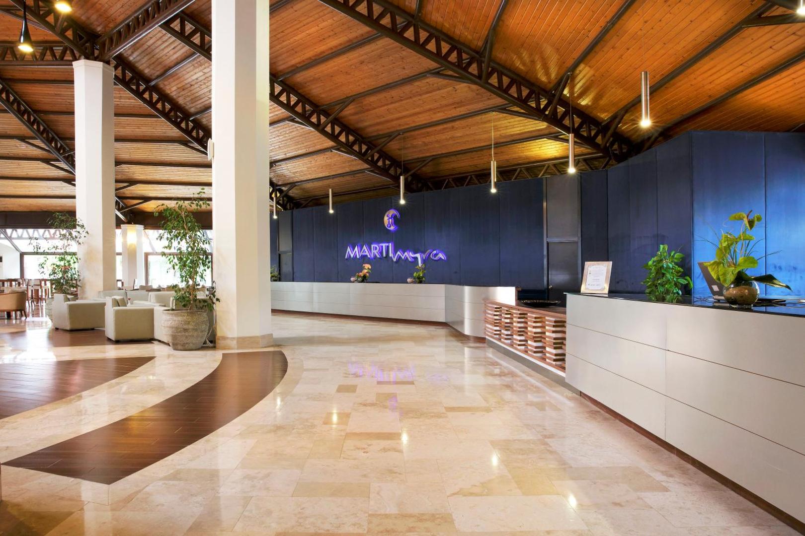 Marti Myra Hotel recepcijas zona ar modernu leti un zilu sienu — gaišs vestibils ar marmora grīdu un koka elementiem