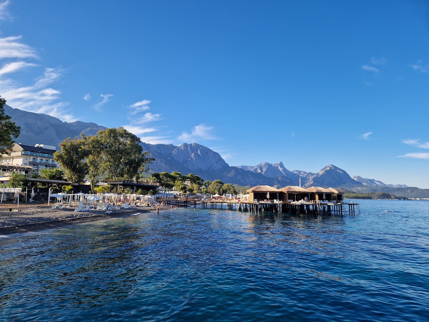 Akra Kemer viesnīcas teritorija no jūras puses — pludmale, koka piestātne ar bungalo un kalnu grēda fonā saulainā dienā
