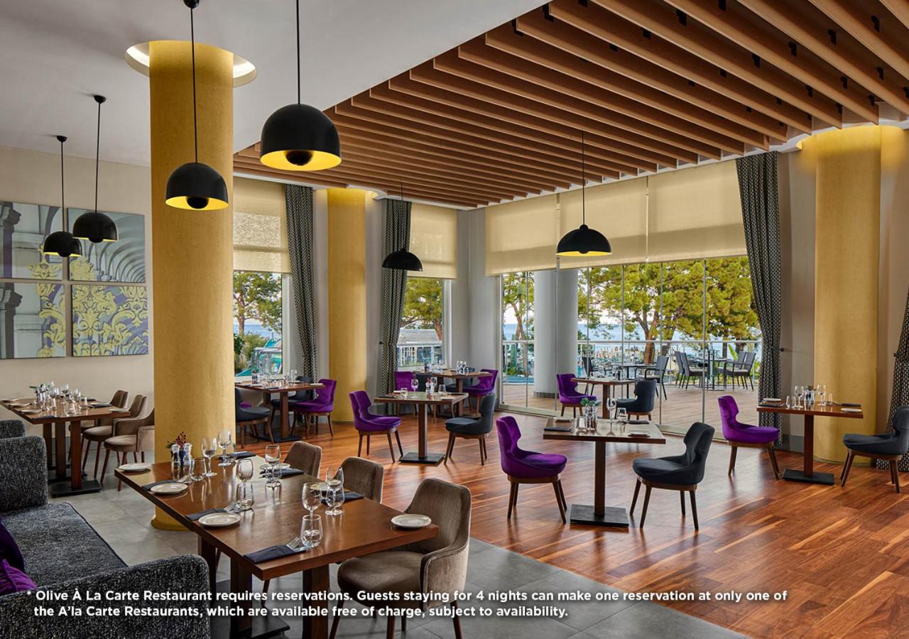 Akra Kemer Olive à la carte restorāna interjers ar modernām mēbelēm, violetiem krēsliem un lieliem logiem uz terasi