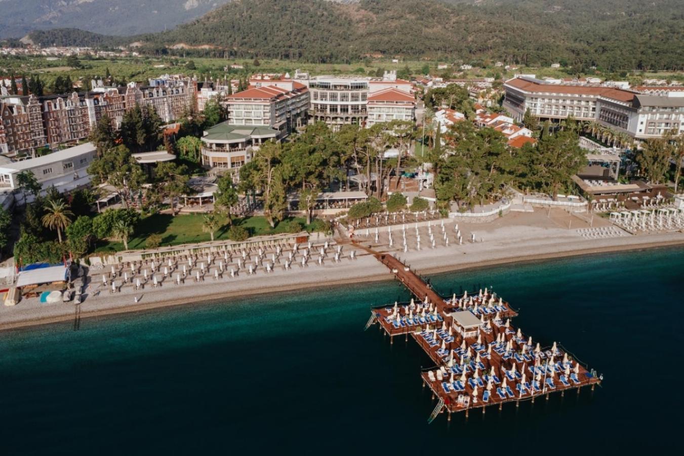 Akra Kemer viesnīcas panorāmas skats no putna lidojuma — privātā pludmale, koka piestātne ar sauļošanās krēsliem un priežu dārzs