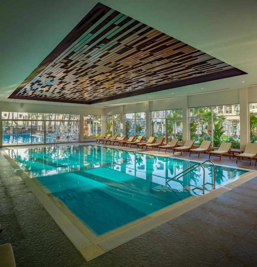Akra Kemer viesnīcas iekštelpu baseins ar panorāmas logiem, atpūtas krēsliem un dekoratīviem griestu elementiem Kemeras reģionā, Turcijā