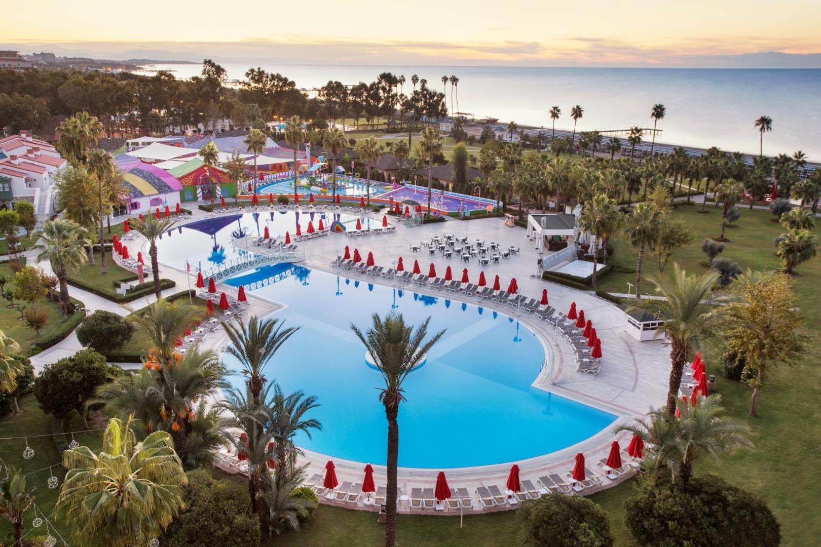 IC Hotels Santai Family Resort plašā teritorija ar lielu āra baseinu, palmām un skatu uz jūru saulrietā Belekā, Turcijā