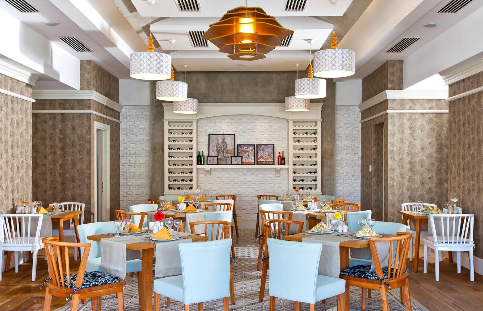 Grand Park Lara viesnīcas a la carte restorāns ar elegantiem galdiem, gaiši ziliem krēsliem un dekoratīvu vīna plauktu pie sienas