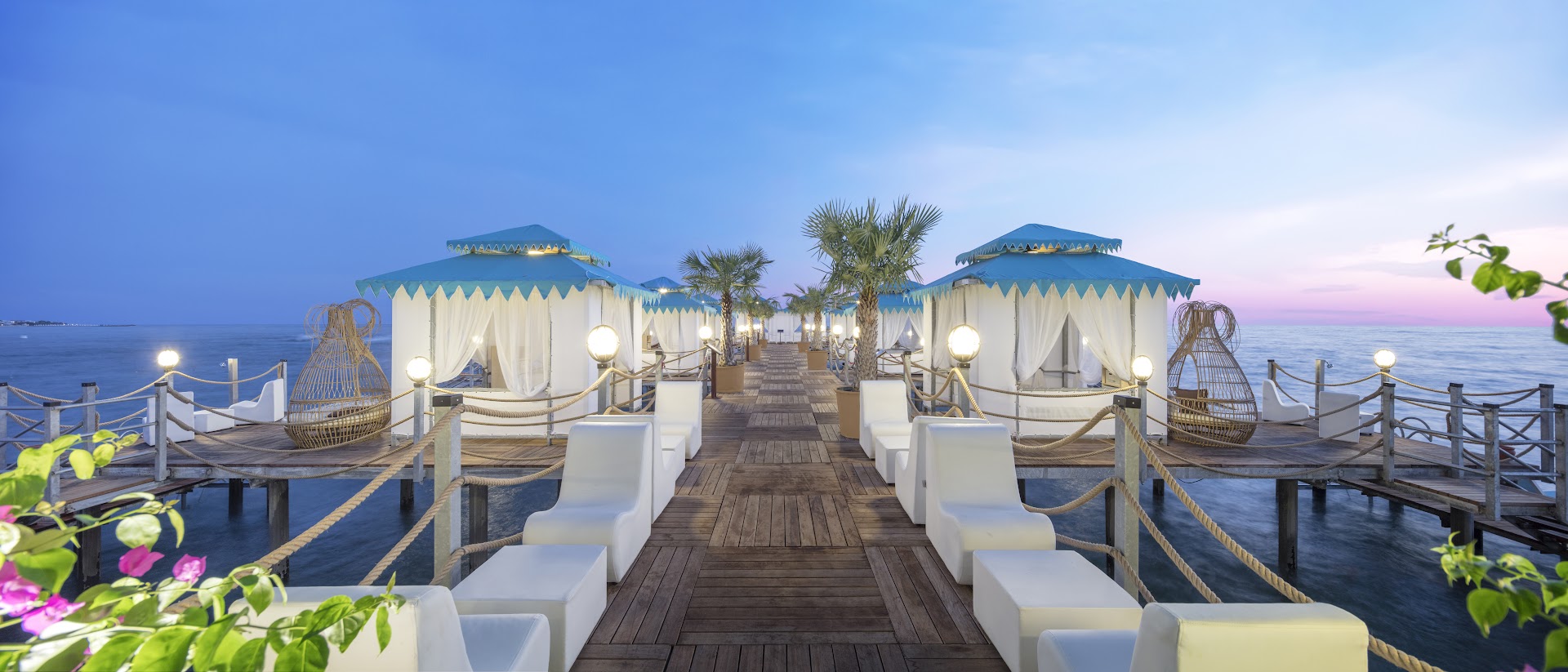 Granada Luxury Beach koka mols jūrā ar privātām atpūtas teltīm, baltiem dīvāniem un palmām saulrieta laikā