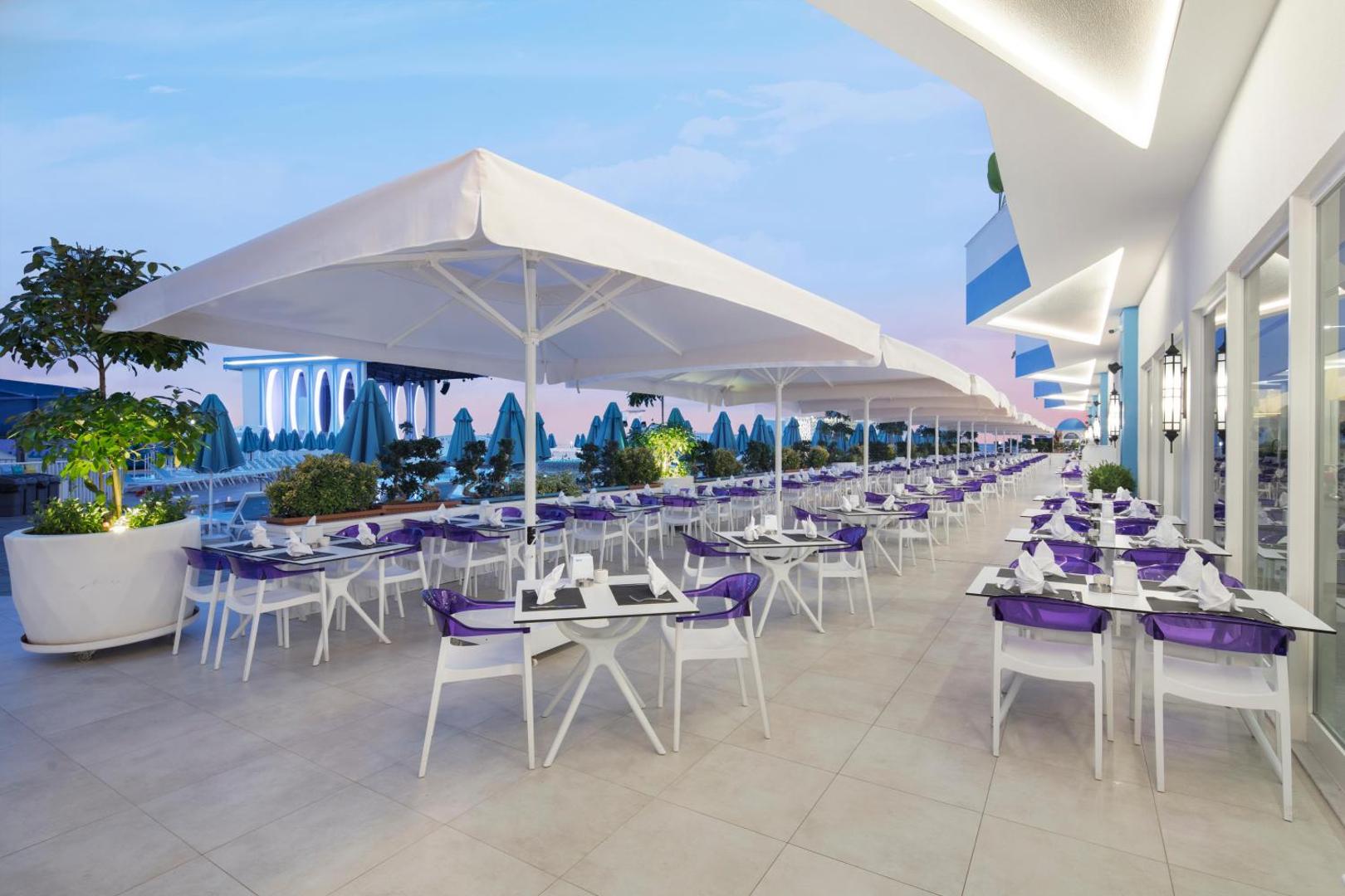 Granada Luxury Beach vakara terase ar balti violetiem krēsliem, lieliem saulessargiem un skatu uz izgaismoto piestātni