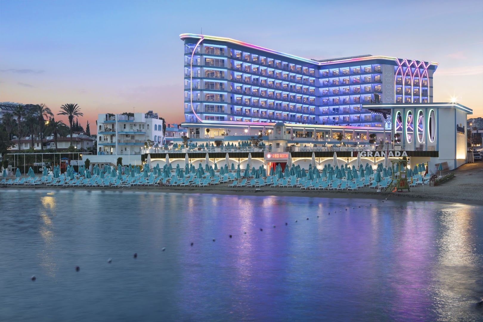 Granada Luxury Beach viesnīcas ēka vakarā ar krāsainu neona apgaismojumu, skats no jūras puses uz pludmali un saulrietu Alanjā