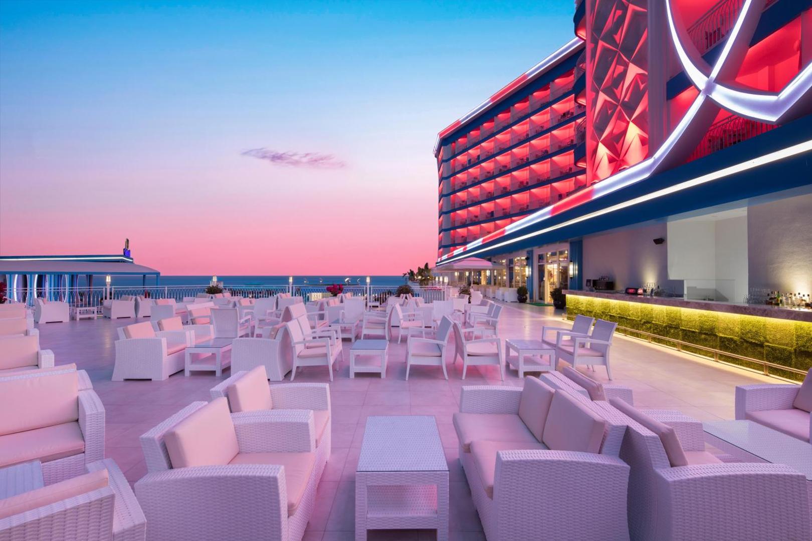Granada Luxury Beach āra terase saulrietā ar baltām mēbelēm un skatu uz Vidusjūru — apgaismota viesnīcas fasāde rozā un zilos toņos