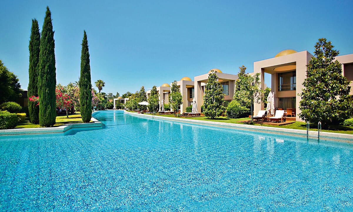 Gloria Serenity Resort villas ar tiešu piekļuvi baseinam, ko ieskauj cipreses un ziedoši dārzi saulainā dienā