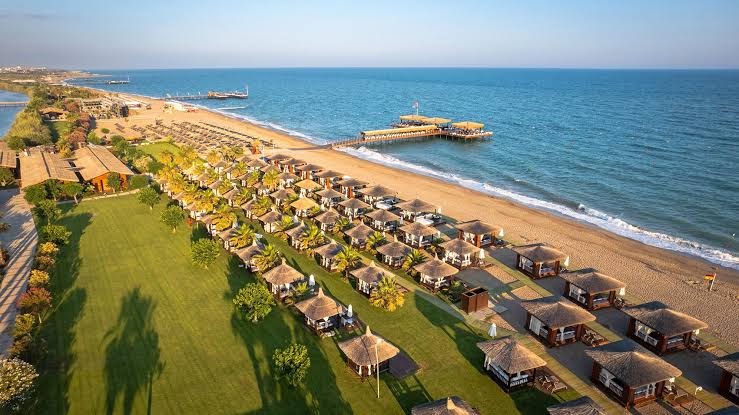 Gloria Serenity Resort panorāmas skats uz plašo pludmali, bungalo zonu un piestātni Belekā saulrieta gaismā