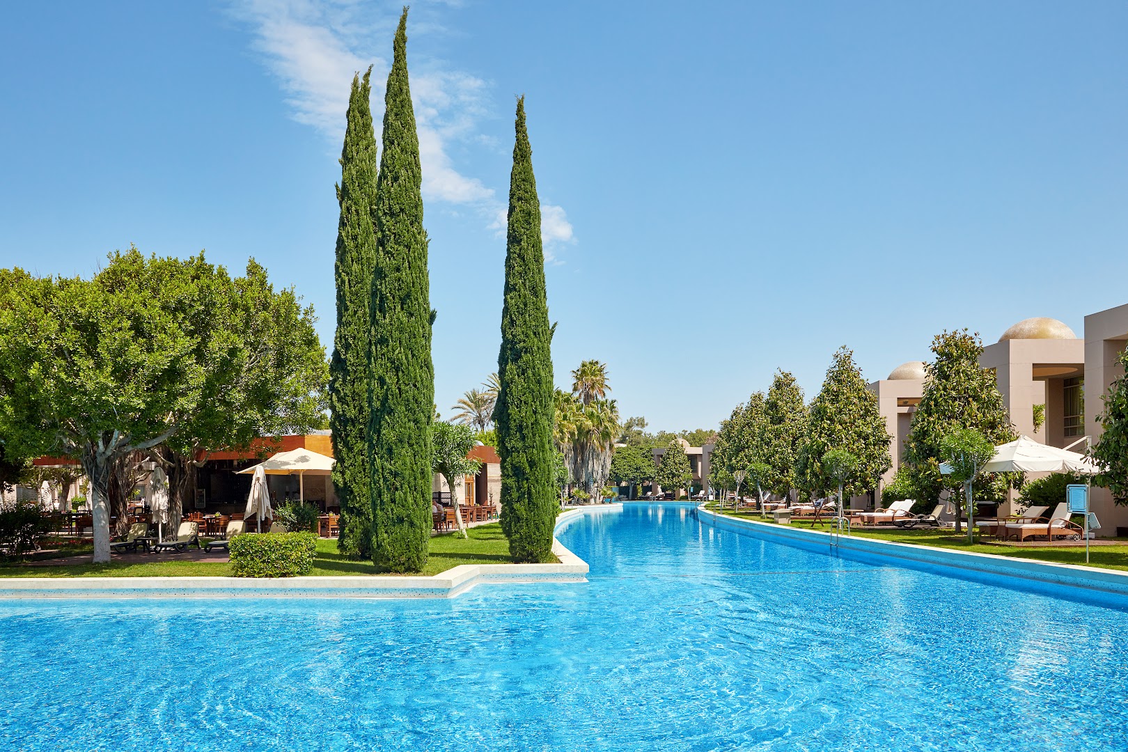 Gloria Serenity Resort dārza skats ar garu peldbaseinu, augstām cipresēm un āra terasi restorāna zonā
