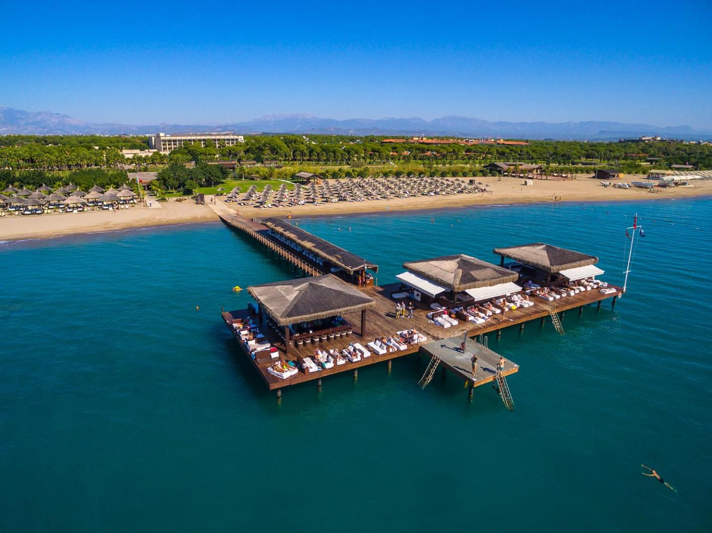 Gloria Serenity Resort privātā pludmale ar plašu koka piestātni, bāru un sauļošanās zonām virs tirkīzzila ūdens Belekā