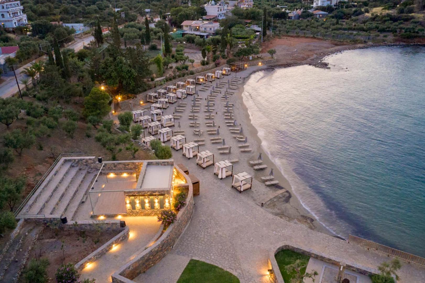 Zeus Eleva Mirabello Bay skats no augšas uz privāto pludmali krēslas stundā — labiekārtota atpūtas zona ar saulessargiem un gultām pie jūras.