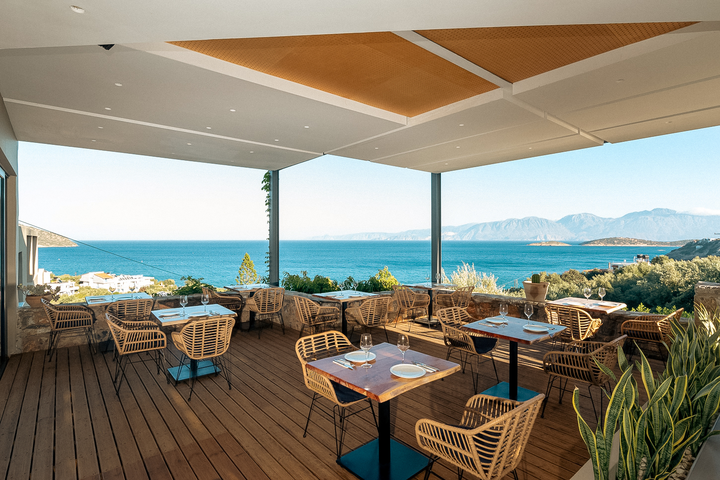 Zeus Eleva Mirabello Bay modernā restorāna terase ar pītiem krēsliem un panorāmas skatu uz zilo jūru un Krētas kalniem