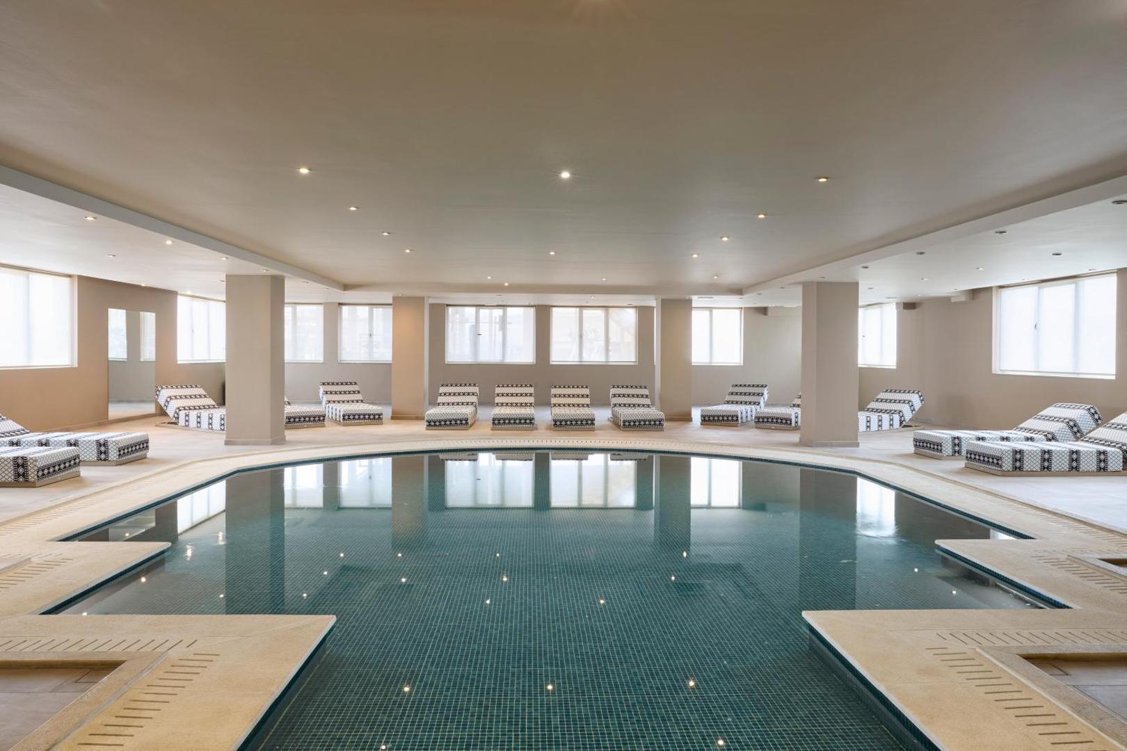Viesnīcas Zeus Eleva Mirabello Bay iekštelpu baseins ar gaišu apdari un ērtiem atpūtas krēsliem relaksācijai spa centrā.