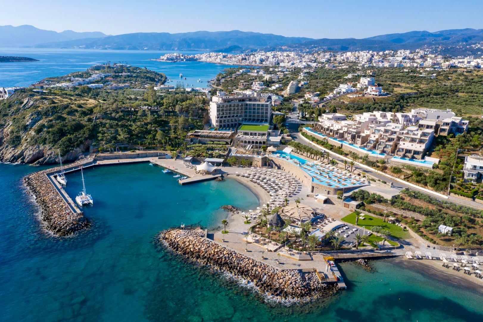 Zeus Eleva Mirabello Bay kūrorta kopskats ar baseiniem, pludmales zonu un apkārtējo piekrastes ainavu saulainā dienā Krētā