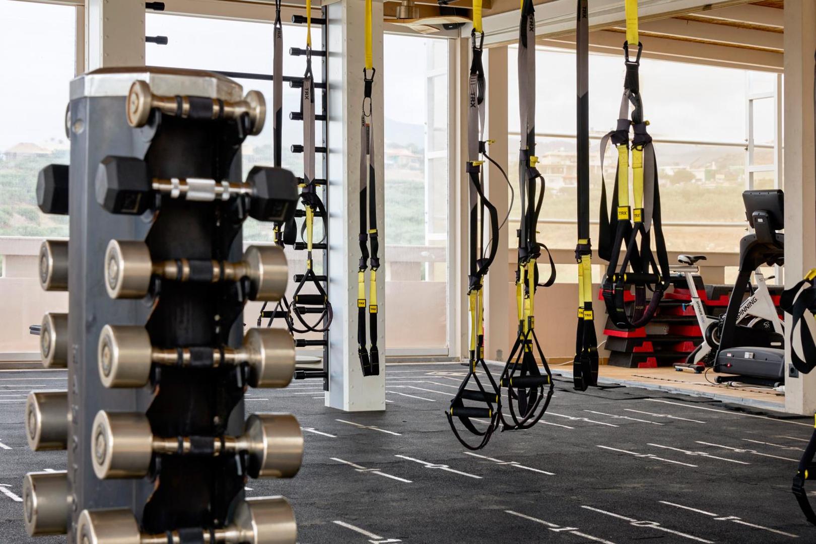 Zeus Eleva Mirabello Bay fitnesa centrs ar TRX piekares sistēmām un hanteļu statīvu — moderna sporta zāle ar skatu uz apkārtni
