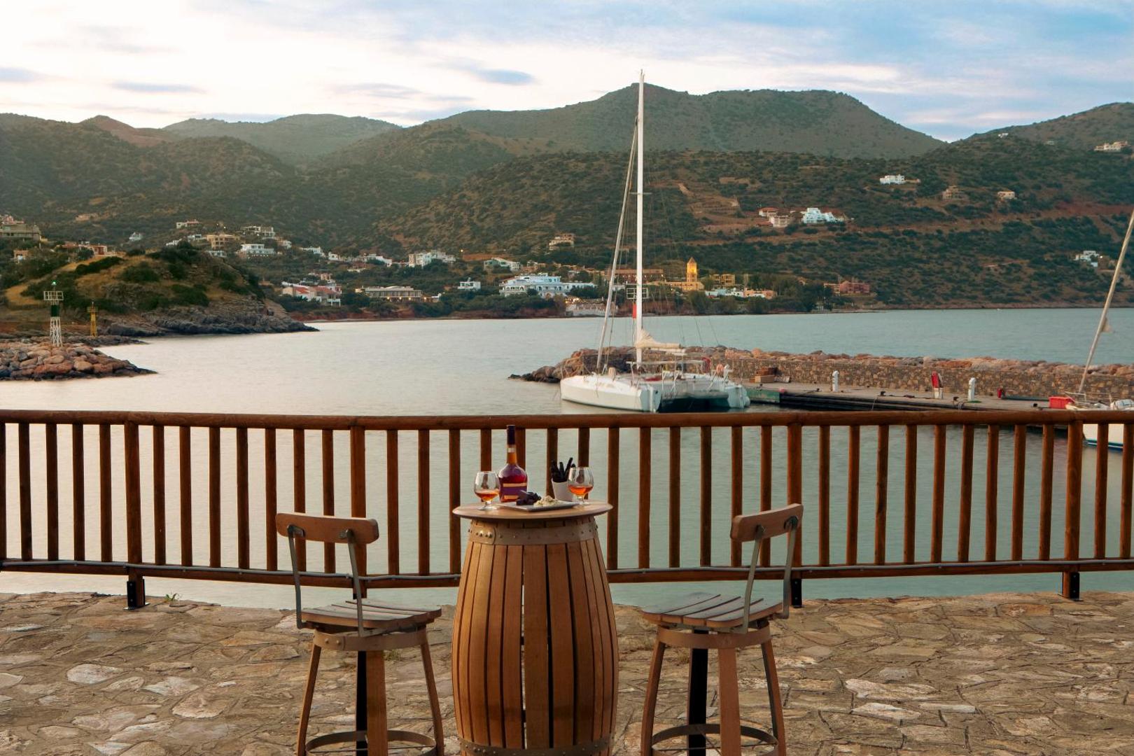 Skats no Zeus Eleva Mirabello Bay terases uz jahtu piestātni un kalniem — galds diviem romantiskam vakaram pie jūras.