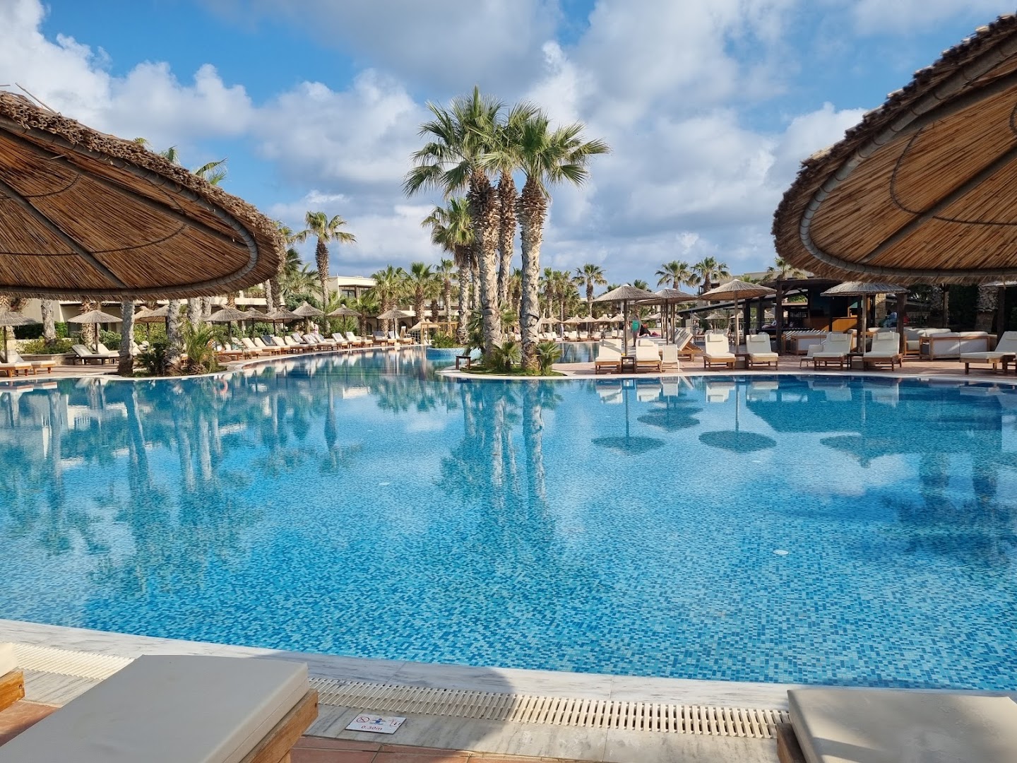 Skats uz Stella Palace Aqua Park Resort baseinu no sauļošanās zonas ar salmu saulessargiem un palmām