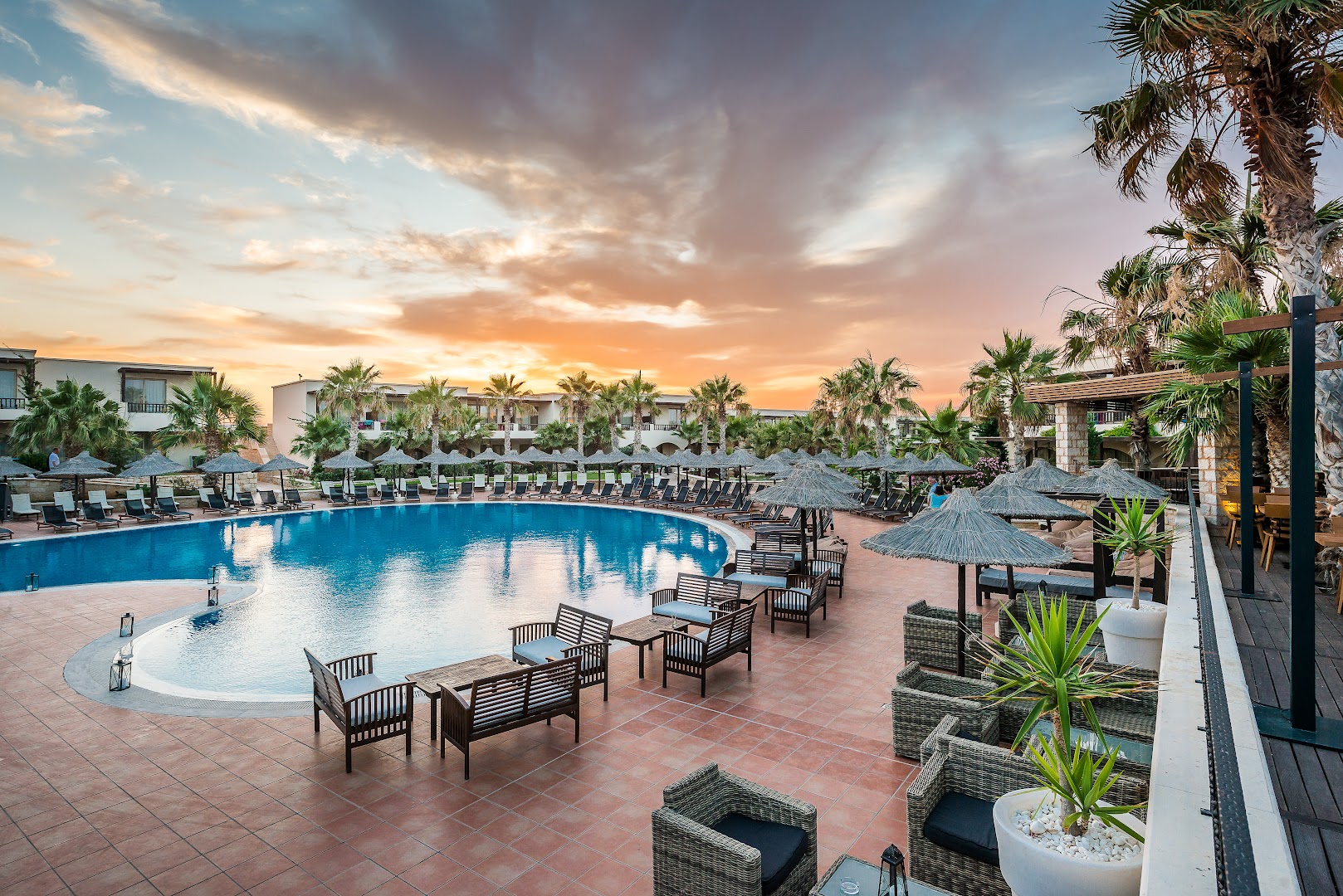 Stella Palace Aqua Park Resort galvenais baseins saulrietā Krētā ar atpūtas krēsliem, saulessargiem un bāra zonu palmu ielenkumā