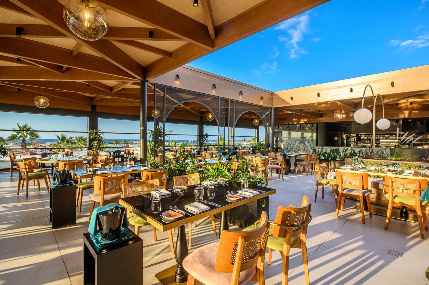 Stella Palace Aqua Park Resort plašā restorāna terase ar koka griestu sijām, modernām mēbelēm un skatu uz jūru Krētā