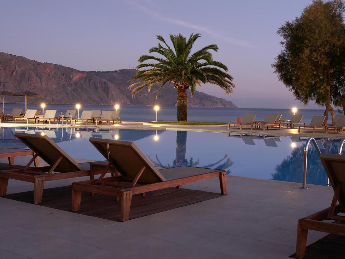 Pilot Beach Resort & Spa āra baseins vakarā ar izgaismotām palmām, sauļošanās krēsliem un skatu uz kalniem Krētā