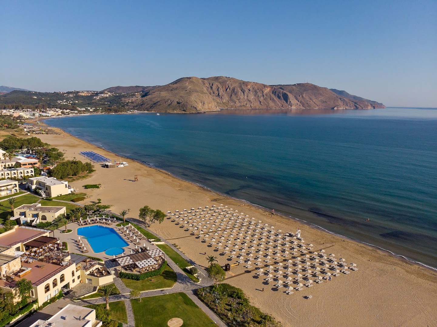 Pilot Beach Resort plašā smilšu pludmale ar daudziem saulessargiem, zilu baseinu un skatu uz līci un kalniem tālumā