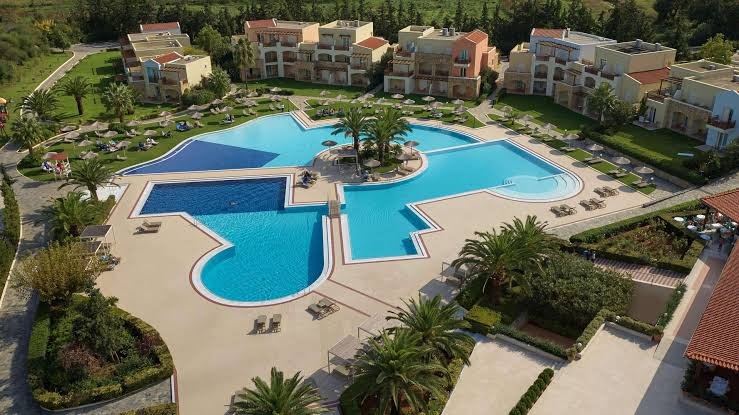 Pilot Beach Resort & Spa viesnīcas teritorijas aerofoto ar unikālas formas baseinu kompleksu, palmām un zemu apbūvi Krētas piekrastē