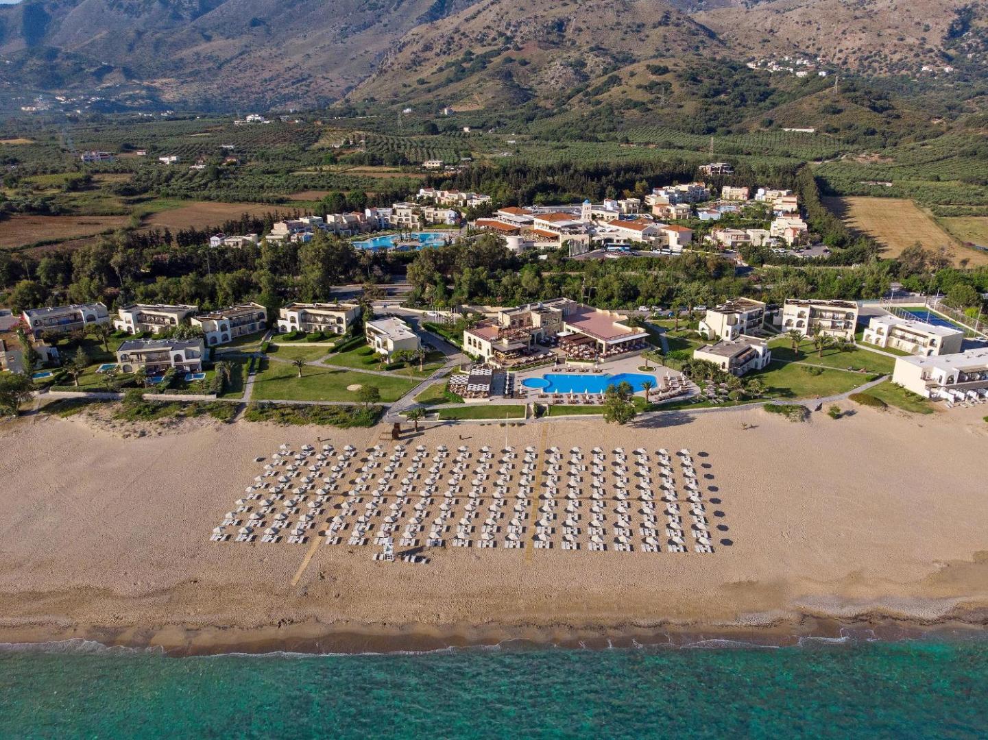 Pilot Beach Resort panorāmas skats no putna lidojuma uz plašo smilšu pludmali, āra baseiniem un kalnu fonu Georgiupoli, Krētā