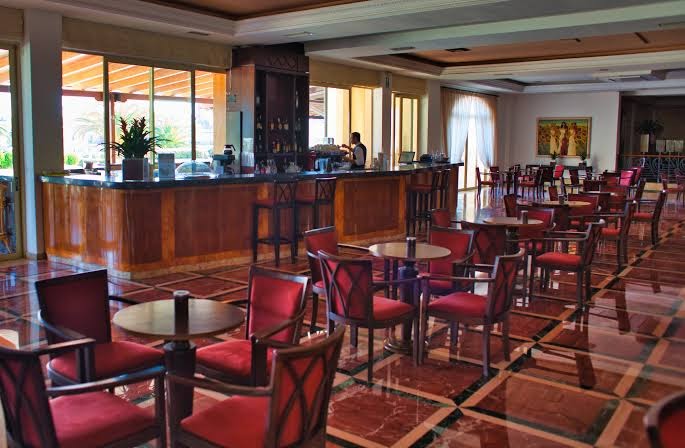 Pilot Beach Resort lobby bārs ar koka apdari un sarkaniem atpūtas krēsliem Georgiupoli, Krētā — plaša telpa ar galdiņiem un bāra leti