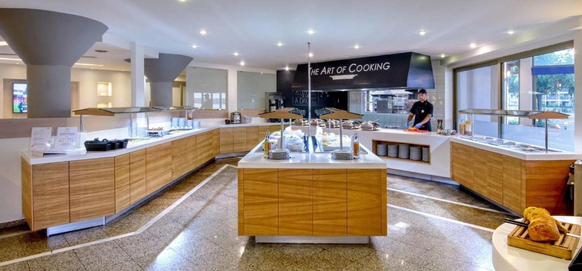 Neptuno Beach viesnīcas restorāna bufetes zona ar koka apdari un atvērto virtuvi 'The Art of Cooking' Krētā