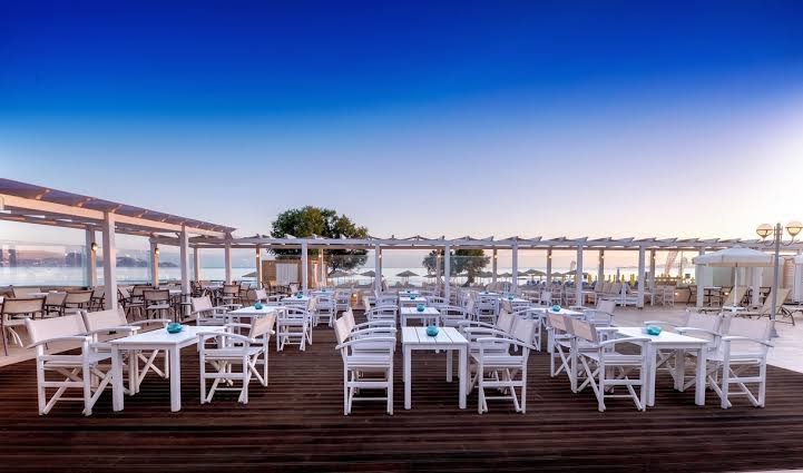 Zeus Hotels Neptuno Beach āra terase ar baltām mēbelēm un koka grīdu Amudarā, Krētā — skats uz jūru saulrietā