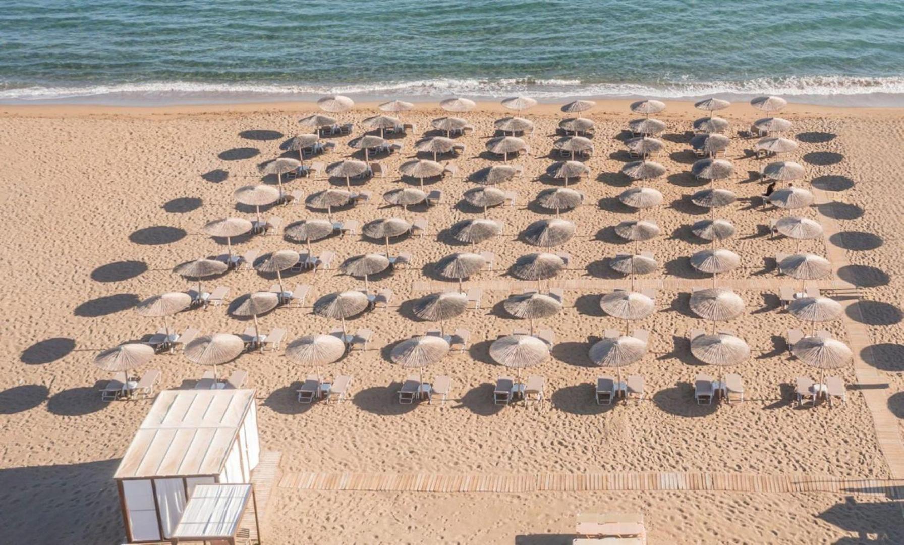 Skats no augšas uz Neptuno Beach privāto smilšu pludmali ar sakārtotām saulessargu rindām un koka laipām Krētā