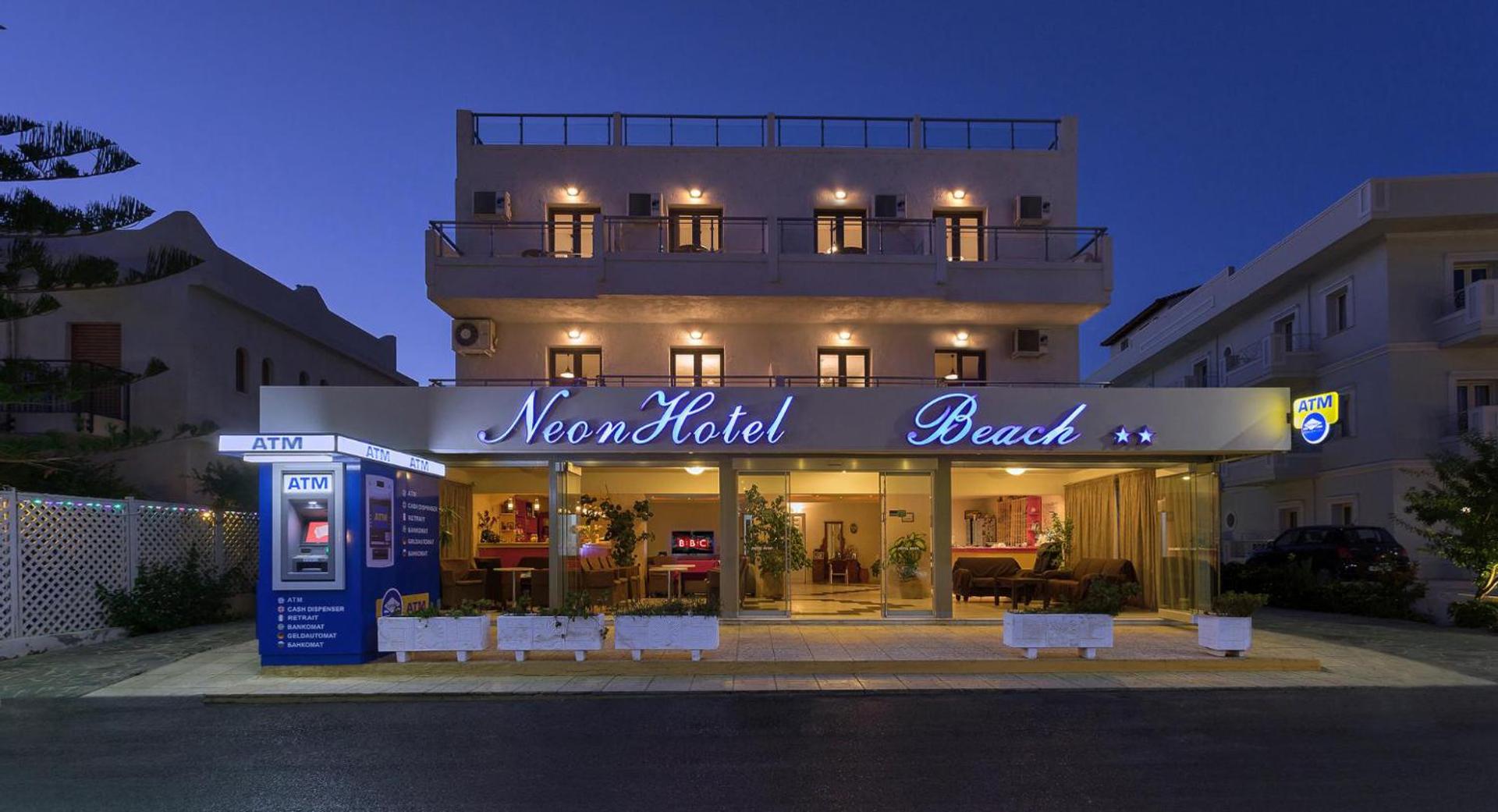 Neon Hotel Beach ieeja vakarā ar izgaismotu izkārtni, bankomātiem un lobby zonu — skats no ielas puses Stalidā