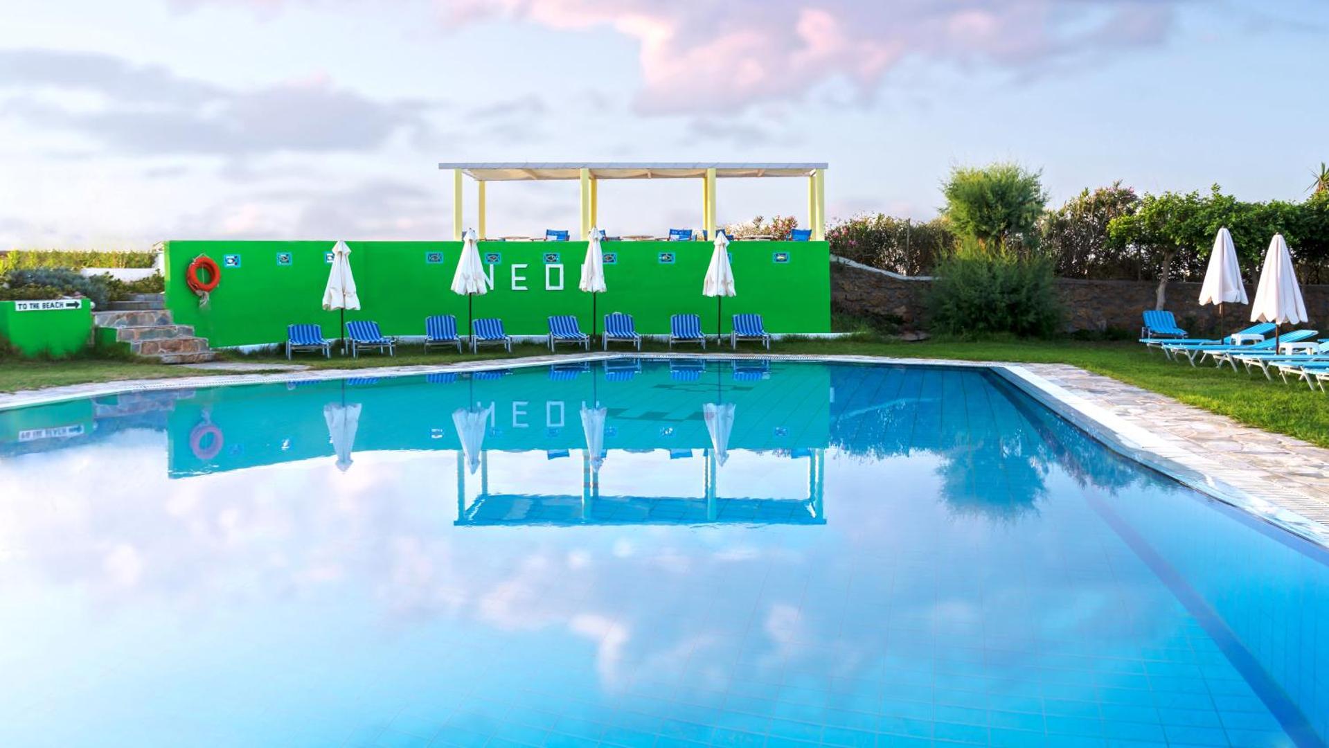 Neon Hotel āra baseins Stalidā, Krētā — zili svītraini sauļošanās krēsli un saulessargi pie koši zaļas dekoratīvās sienas vakara gaismā