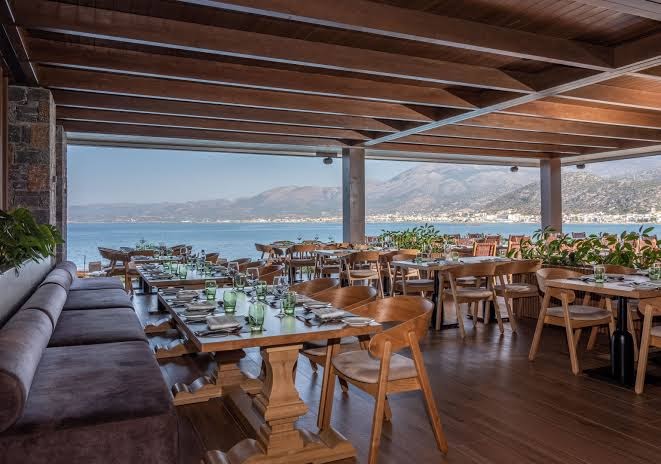 Nana Golden Beach restorāns ar koka apdari un panorāmas skatu uz jūru un kalniem Krētas piekrastē saulainā dienā